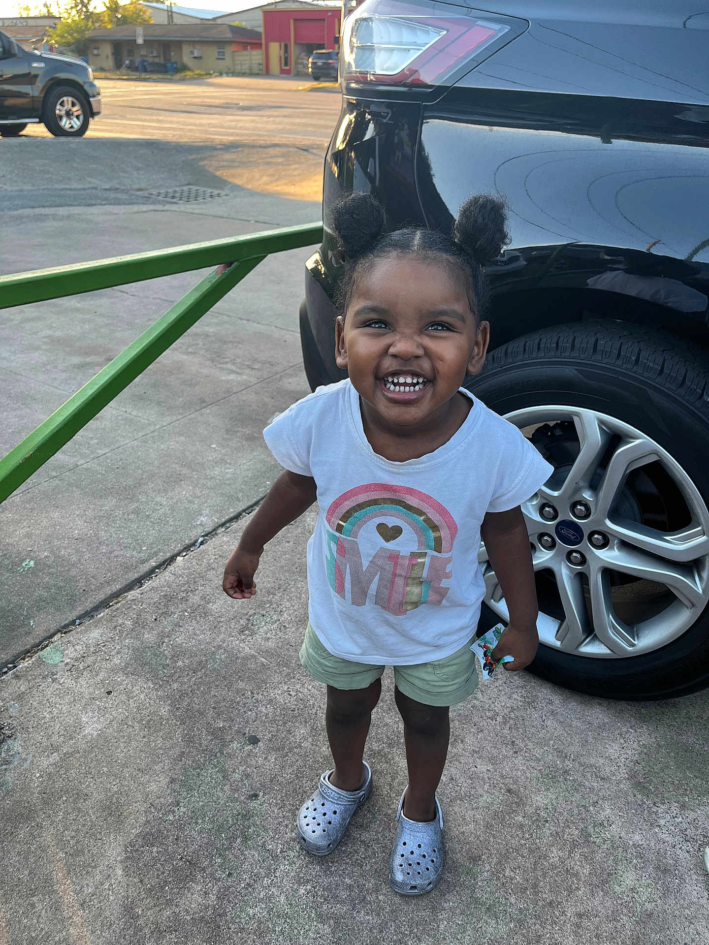 Naleighna is registered to the contest to win money with this photo: toddler, child, smile, hair_buns, tshirt, shorts, crocs, car, tire, concrete, outdoor, daylight, happy, person, standing, joyful, vehicle, sidewalk, casual_clothing, sunlight