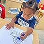 child, toddler, drawing, coloring, marker, table, classroom, hair_buns, blue_marker, paper, furniture, floor, indoor, preschool, education, kid, artwork, concentration, clothing, person