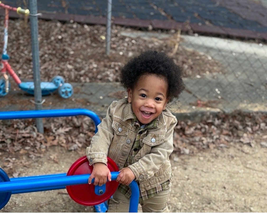 Amiel joined the competition — help win amazing prizes! child, toddler, smiling, playground, play_equipment, outdoor, happy, afro, jacket, denim_jacket, red, blue, fence, leaves, dirt, person, casual_clothing, young_child, daylight, park