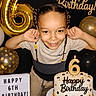 balloons, birthday, birthday_cake, birthday_sign, black_decorations, braids, cake, candles, celebration, child, confetti, flexing, gold_decorations, happy, indoors, number_six, party, portrait, six_years_old, smiling