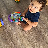 Edem a rejoint le concours — aidez-le/la à gagner de superbes lots ! barefoot, blue_clothing, book, child, colorful, curly_hair, expression, face, floor, hand, home, indoor, looking_up, playful, plush_toy, pouf, sitting, toddler, toy, wooden_floor