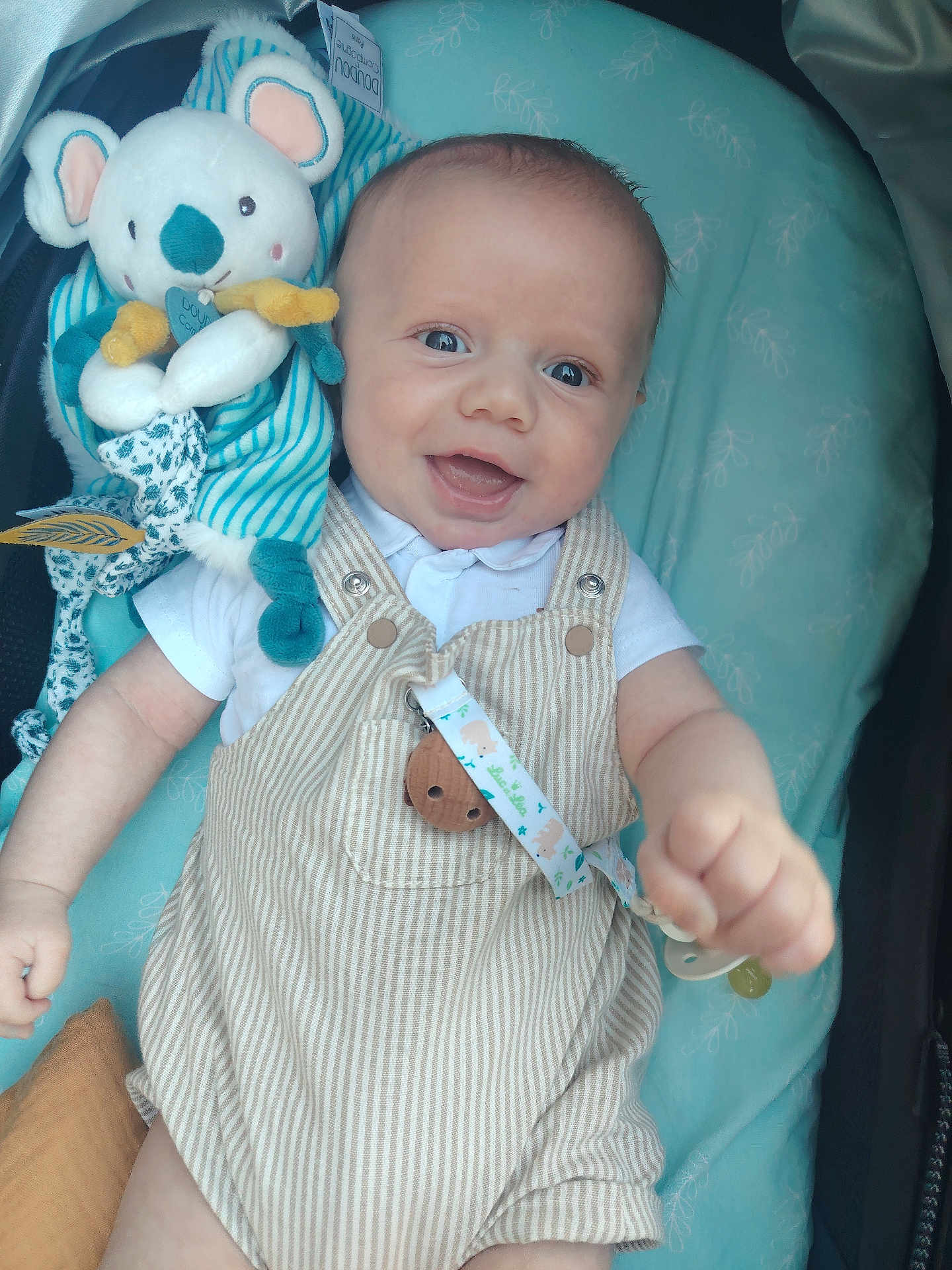Soann a rejoint le concours — aidez-le/la à gagner de superbes lots ! baby, smiling, plush_toy, koala, striped_overalls, white_shirt, lying_down, blue_background, happy, infant, child, cute, face, hand, pacifier_clip, button, fabric, soft_toy, closeup, indoors