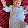 baby, infant, onesie, white_clothing, smiling, happy, lying_down, blanket, striped_pattern, hand, face, cheerful, cute, child, young, soft_fabric, bed, indoors, person, portrait