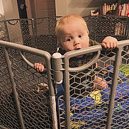 Lennox is registered to the contest to win money with this photo: baby, infant, child, playpen, baby_gate, mesh, hands, grip, curious, blue_blanket, toys, indoor, bookshelf, home, sitting, portrait, wide_eyes, mouth_open, tshirt, floor_mat
