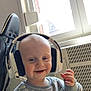 toddler, headphones, smiling, indoor, window, sweater, dinosaur_pattern, child, happy, face, person, clothing, radiator, chair, black_chair, toy, hand, light, cozy, casual