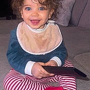 Meylie participe au concours pour gagner de l'argent avec cette photo : toddler, child, curly_hair, blue_eyes, bib, striped_pants, red_socks, remote_control, smiling, sitting, couch, indoor, happy, baby, cute, person, portrait, cozy, clothing, home