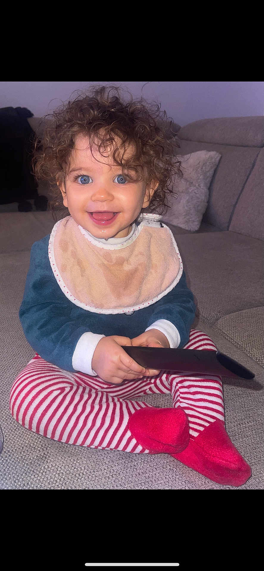 Meylie participe au concours pour gagner de l'argent avec cette photo : toddler, child, curly_hair, blue_eyes, bib, striped_pants, red_socks, remote_control, smiling, sitting, couch, indoor, happy, baby, cute, person, portrait, cozy, clothing, home