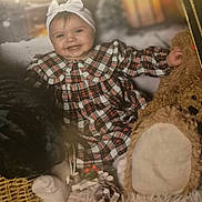 Mylah is registered to the contest to win money with this photo: baby, child, plaid_dress, headband, smiling, teddy_bear, blanket, holiday_decorations, pine_cones, basket, soft_toys, festive, indoor, portrait, cute, happy, cozy, winter_theme, childrens_clothing, sitting