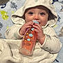 baby, bottle, blanket, hood, infant, cute, smiling, blue_eyes, hand, face, clothing, cozy, child, indoors, soft_texture, patterned_blanket, warm, lying_down, adorable, feeding