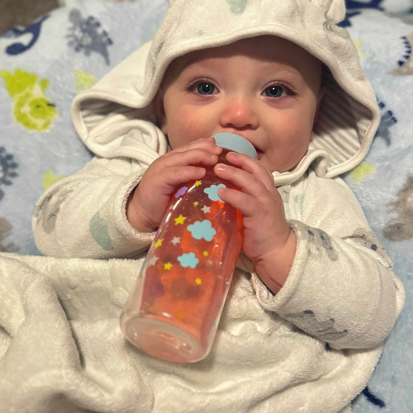 Waylon is registered to the contest to win money with this photo: adorable, baby, blanket, blue_eyes, bottle, child, clothing, cozy, cute, face, feeding, hand, hood, indoors, infant, lying_down, patterned_blanket, smiling, soft_texture, warm