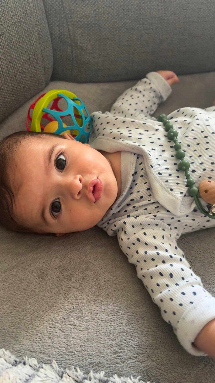 Adib a rejoint le concours — aidez-le/la à gagner de superbes lots ! baby, infant, face, polka_dot, onesie, toy, ball, couch, gray, puckered_lips, wide_eyes, child, soft_texture, indoors, cute, lying_down, head, hand, expression, portrait