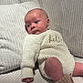 baby, infant, knitted_outfit, cream_color, couch, pillows, soft_furniture, indoor, cute, child, portrait, cozy, sitting, looking_away, warm_lighting, hand, footwear, ribbed_texture, home, relaxed