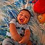 baby, smiling, laughing, child, plush_toy, stuffed_animal, blanket, pattern, bed, blue, white, red, yellow, clothing, onesie, infant, happy, cute, toy, indoor