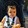child, toddler, stroller, seatbelt, sunlight, outdoor, clothing, white_shirt, hand, face, expression, hair, skin, person, baby, daylight, urban, sidewalk, vehicle, background