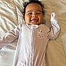baby, bed, cheeky, child, cozy, curly_hair, cute, cute_expression, happy, indoor, infant, lying_down, onesie, portrait, purple_clothing, quilt, smiling, soft_lighting, tongue_out, young_child