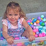 child, girl, smiling, plastic_container, colorful_eggs, easter_eggs, playful, curly_hair, pink_shirt, overalls, indoor, happy, face, hands, toy, pastel_colors, cute, fun, young, person