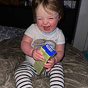Olivia is registered to the contest to win money with this photo: toddler, child, laughing, smiling, striped_pants, white_shirt, indoor, blanket, holding_object, happy, rosy_cheeks, sitting, baby, face, person, young_child, cozy, candid, cute, playful