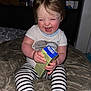 toddler, child, laughing, smiling, striped_pants, white_shirt, indoor, blanket, holding_object, happy, rosy_cheeks, sitting, baby, face, person, young_child, cozy, candid, cute, playful