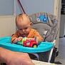baby, blue_tray, bread, child, cooking, cutting, face, family, food, furniture, hand, high_chair, home, indoor, kitchen, orange_shirt, person, playing, table, toy