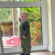 Ronnie joined the competition — help win amazing prizes! toddler, child, indoor, glass_door, overalls, striped_shirt, pointing, curious, greenery, plants, toy, floor, socks, window, natural_light, home, person, young_child, standing, playful