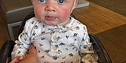 Kenzie is registered to the contest to win money with this photo: baby, child, high_chair, indoor, blue_eyes, hand, white_shirt, patterned_clothing, curious, rosy_cheeks, seated, person, furniture, table, floor, light, skin, young_child, cute, expression