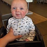 Kenzie joined the competition — help win amazing prizes! baby, child, high_chair, hand, white_shirt, floral_pattern, blue_eyes, smile, indoor, restaurant, table, chair, wooden_floor, person, cute, infant, skin, head, seated, support