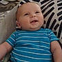 baby, infant, blue_onesie, striped_clothing, smiling, face, head, legs, cushion, patterned_cushion, indoor, floor, wooden_floor, shoes, black_shoes, happy, child, person, seated, home