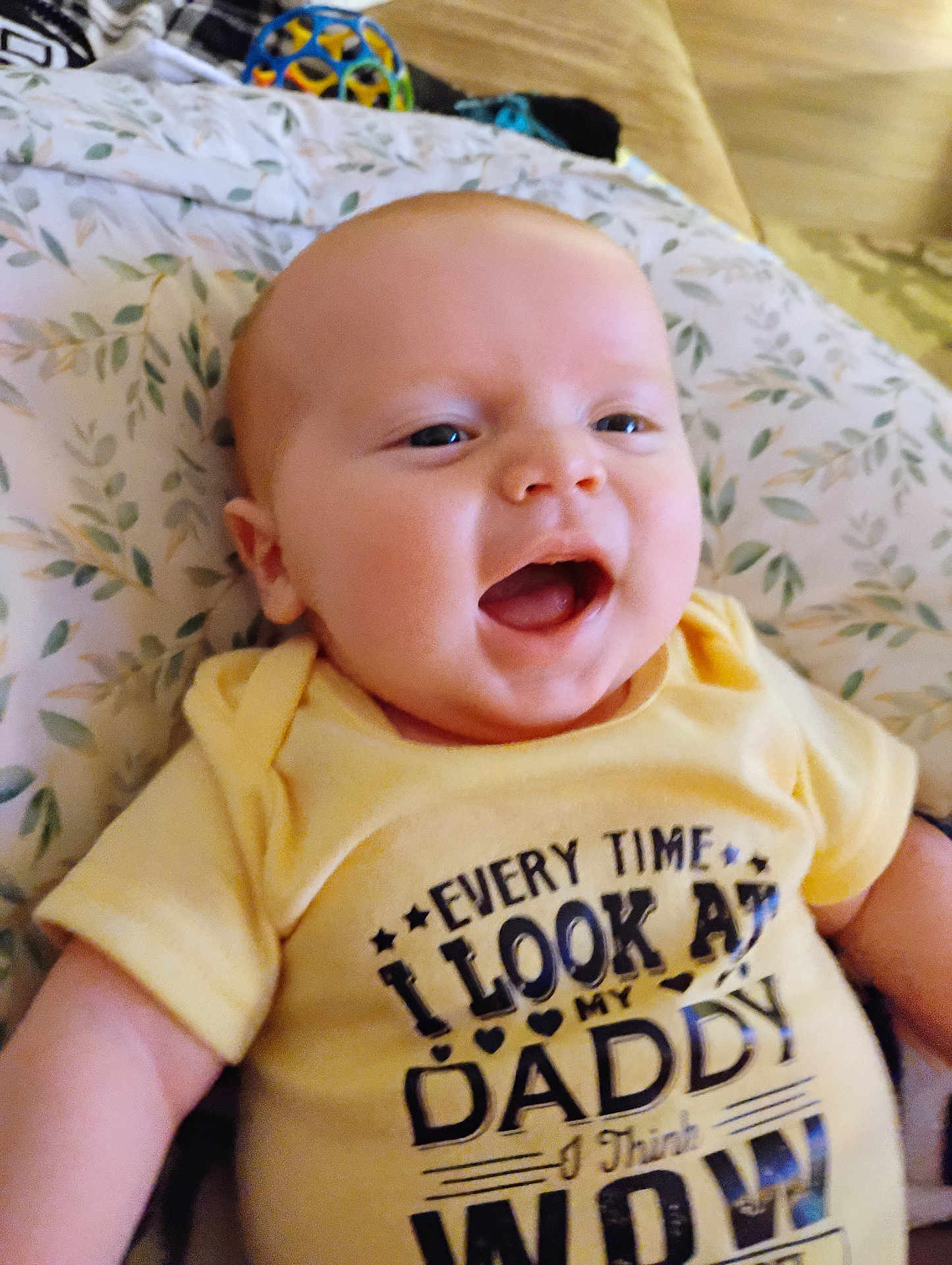 Jace is registered to the contest to win money with this photo: baby, infant, smiling, yellow_shirt, text_on_clothing, pillow, leaf_pattern, happy, child, face, mouth_open, cute, indoors, person, young_child, head, skin, eyes, sleeves, toddler