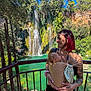 woman, baby, waterfall, nature, outdoor, tattoo, baby_carrier, greenery, blue_sky, smile, person, river, trees, sunlight, wooden_platform, holding_baby, short_hair, fall, scenic, relaxing