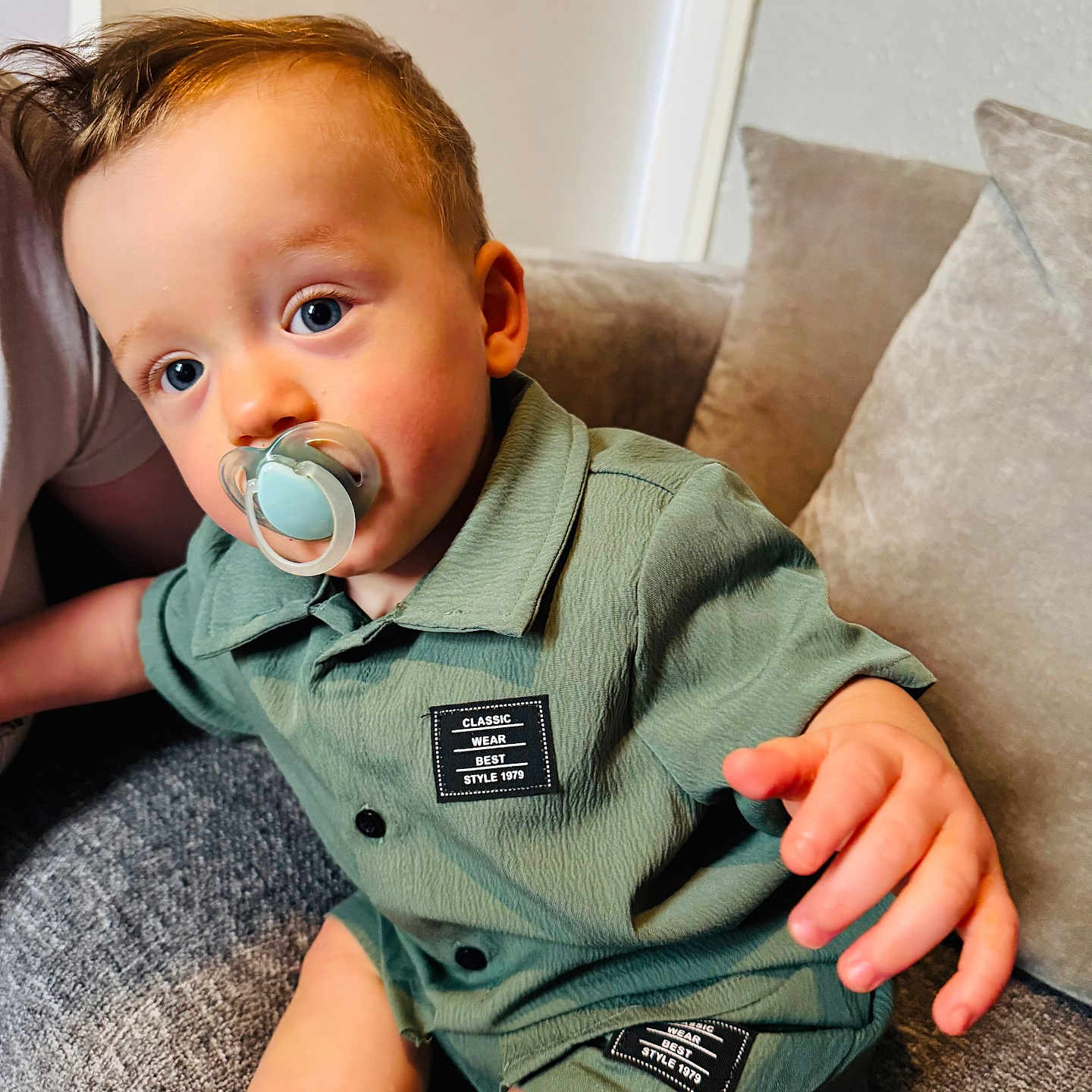 Sonny is registered to the contest to win money with this photo: blue_eyes, casual_clothing, child, comfortable, couch, curly_hair, cute, face, green_outfit, hand, home, indoor, pacifier, person, plush_cushion, shirt, shorts, sitting, toddler, young_child