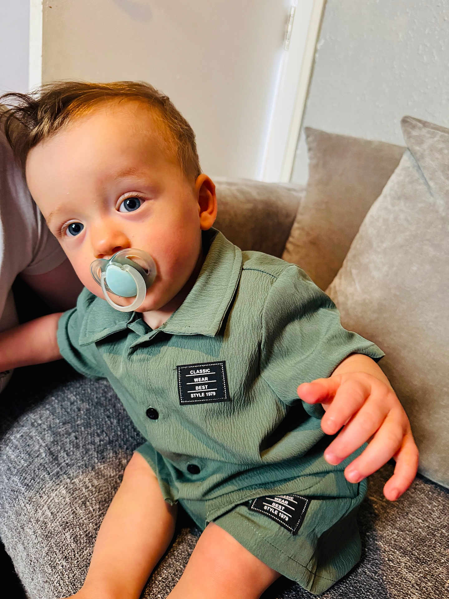 Sonny is registered to the contest to win money with this photo: toddler, child, pacifier, green_outfit, shorts, shirt, blue_eyes, curly_hair, couch, indoor, home, plush_cushion, person, sitting, hand, face, cute, young_child, casual_clothing, comfortable