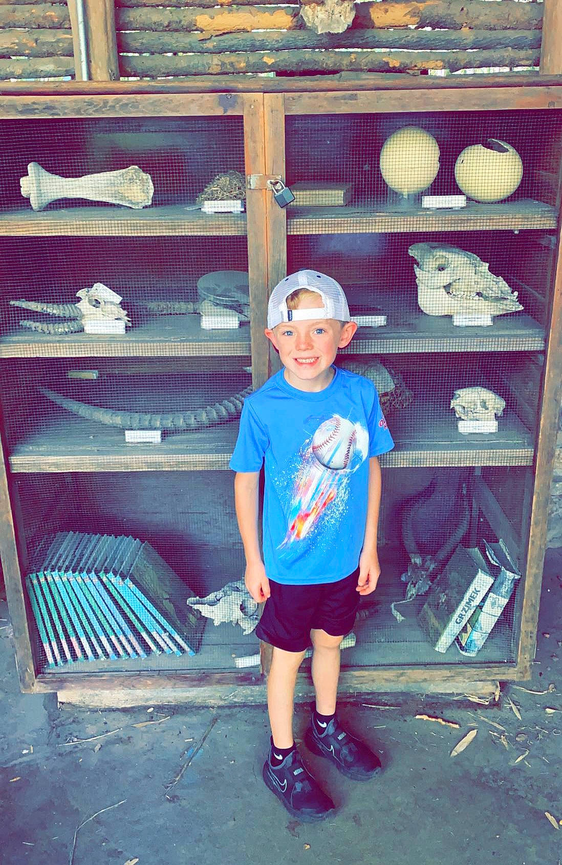Reece is registered to the contest to win money with this photo: baseball_cap, cap, child, electric_blue, eyewear, footwear, happy, hat, headwear, helmet, joy, person, shelf, shelving, shoe, shorts, smile, sneakers, standing, sunglasses