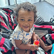 Kai joined the competition — help win amazing prizes! baby, child, smiling, curly_hair, sitting, toy, colorful_toy, bed, bedroom, blanket, striped_bedding, portrait, indoors, happy, infant, clothing, short_sleeves, barefoot, playtime, photography