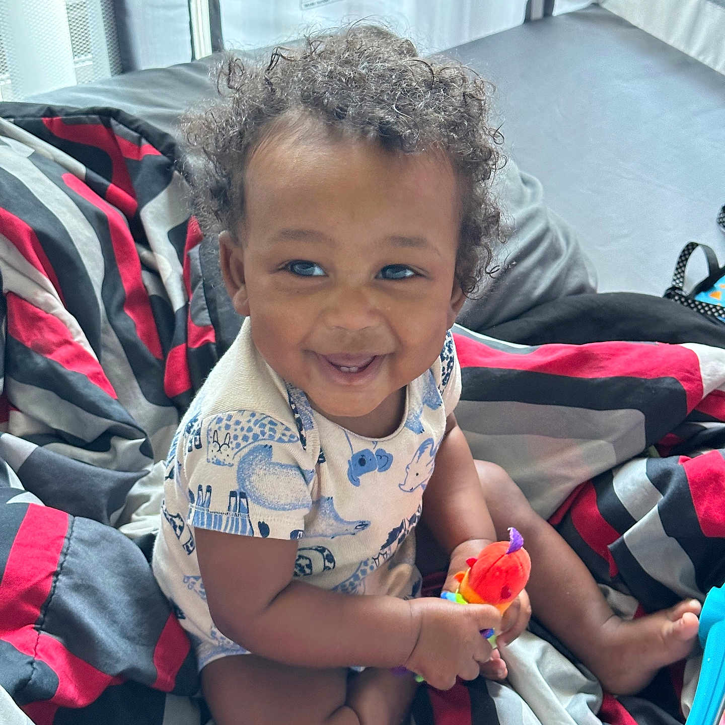 Kai joined the competition — help win amazing prizes! baby, child, smiling, curly_hair, sitting, toy, colorful_toy, bed, bedroom, blanket, striped_bedding, portrait, indoors, happy, infant, clothing, short_sleeves, barefoot, playtime, photography