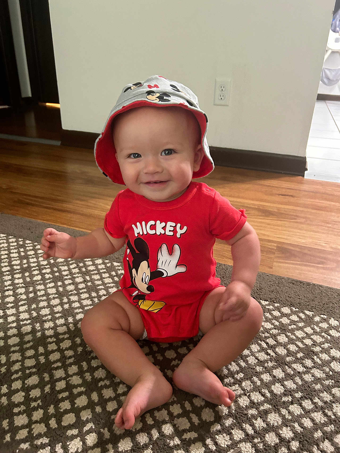 Karson is registered to the contest to win money with this photo: baby, child, mickey_mouse, red_clothing, hat, bucket_hat, smiling, sitting, carpet, floor, wooden_floor, indoor, cute, infant, person, face, happy, toddler, patterned_carpet, onesie
