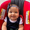 baby, infant, child, smiling, portrait, headband, bow, car_seat, red, black, sitting, indoors, face, eyes, hands, clothing, dress, safety_label, close_up, happy