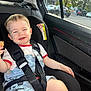 Lucian is registered to the contest to win money with this photo: toddler, child, car_seat, seat_belt, snack, smiling, mischievous, car_interior, window, daylight, clothing, shorts, tshirt, mickey_mouse, hair, face, hand, safety, vehicle, parking_lot