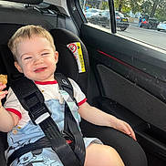 Lucian is registered to the contest to win money with this photo: toddler, child, car_seat, seat_belt, snack, smiling, mischievous, car_interior, window, daylight, clothing, shorts, tshirt, mickey_mouse, hair, face, hand, safety, vehicle, parking_lot