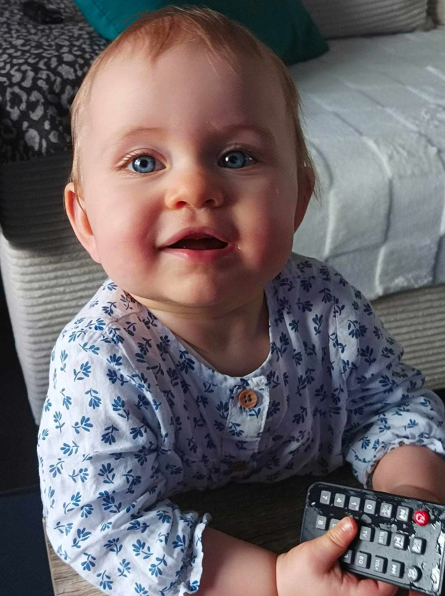 Victoire participe au concours pour gagner de l'argent avec cette photo : baby, infant, child, blue_eyes, smiling, face, cheeks, patterned_shirt, floral_print, remote_control, hand, wooden_table, couch, cushion, indoor, portrait, curious, blonde_hair, closeup, cozy