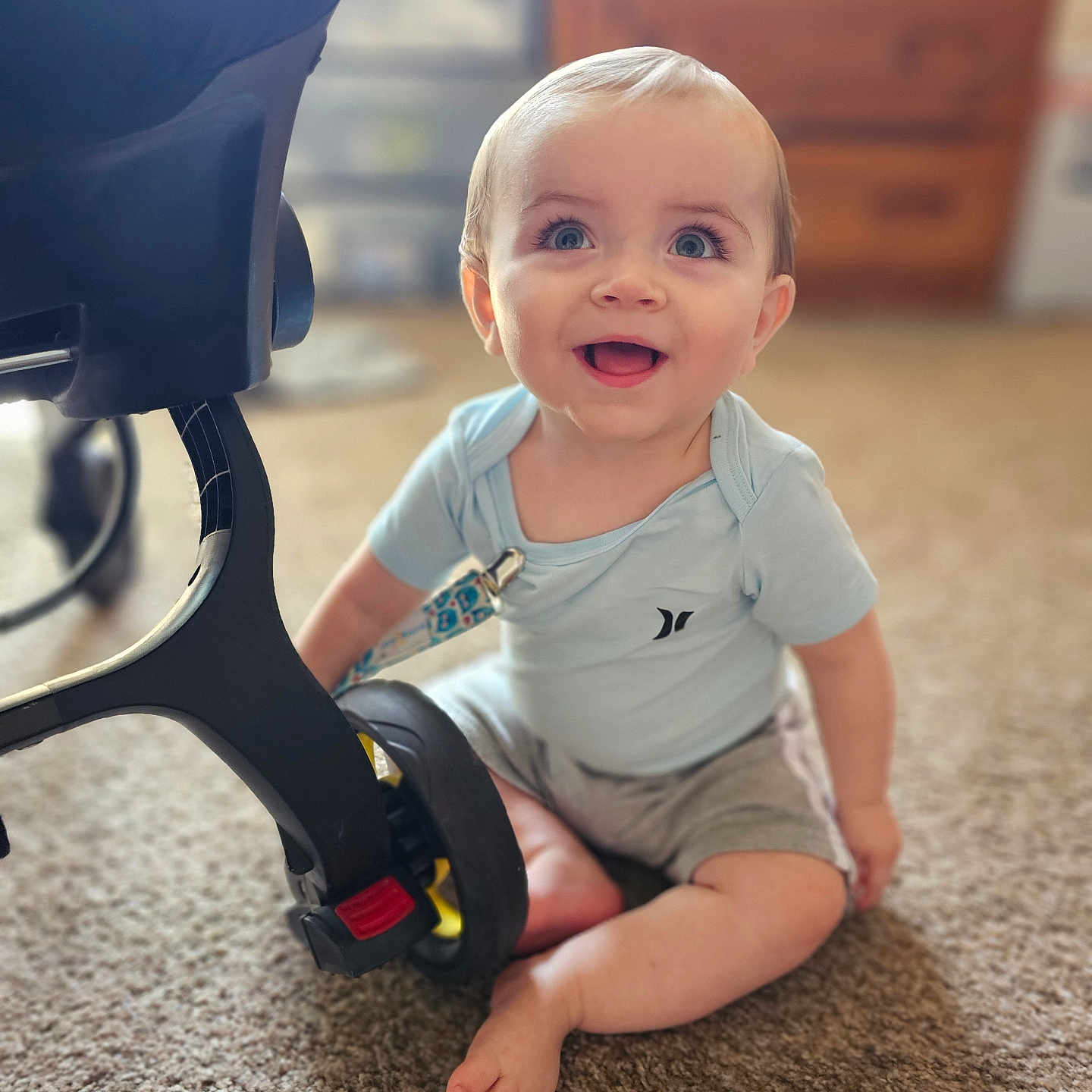 Keegan is registered to the contest to win money with this photo: baby, bodypart, clothing, e_scooter, face, finger, hand, happy, head, indoors, pants, person, photography, portrait, shorts, sitting, t_shirt, transportation, vehicle, wood