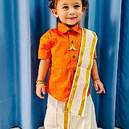 Shlok is registered to the contest to win money with this photo: blue_curtain, bracelets, child, cultural, curly_hair, cute, fashion, gold_border, happy, indoor, necklace, orange_shirt, person, portrait, smiling, standing, toddler, traditional_clothing, white_dhoti, young_child