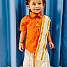 blue_curtain, bracelets, child, cultural, curly_hair, cute, fashion, gold_border, happy, indoor, necklace, orange_shirt, person, portrait, smiling, standing, toddler, traditional_clothing, white_dhoti, young_child
