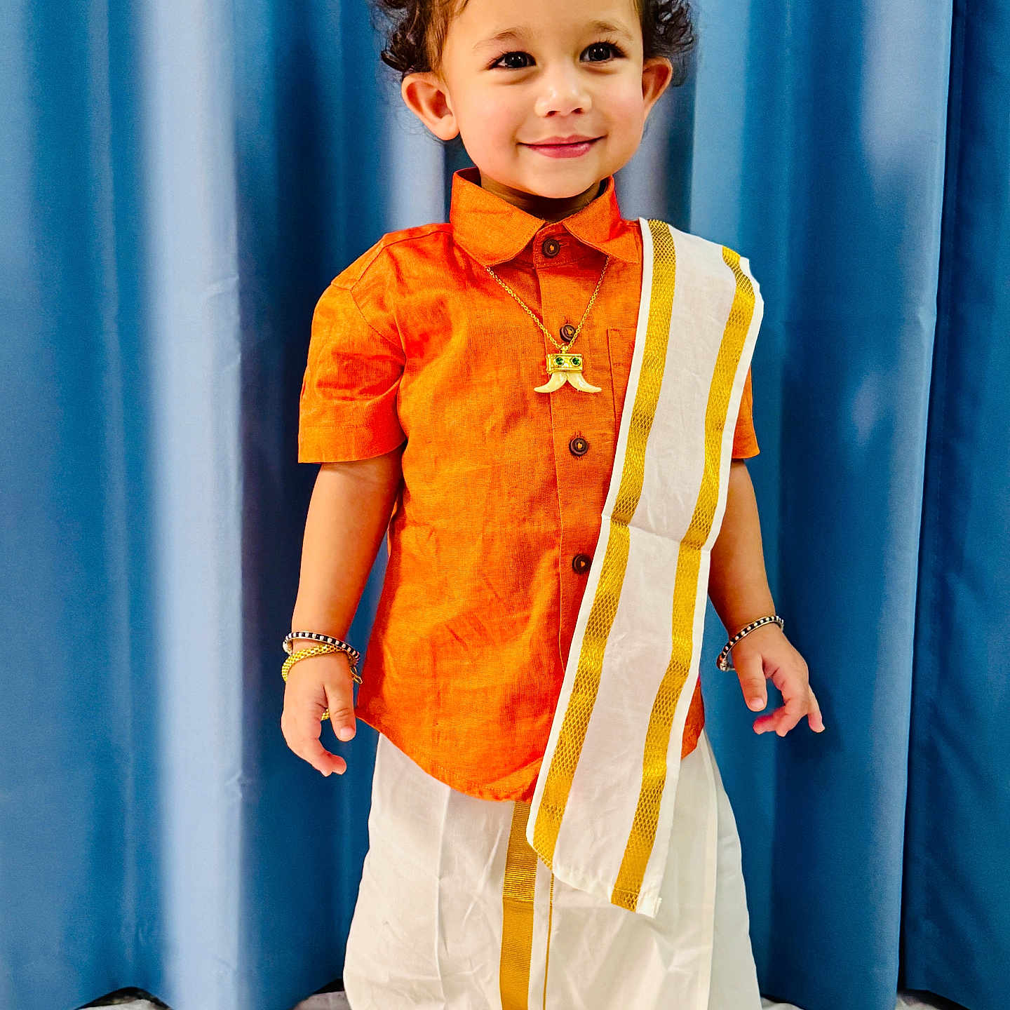 Shlok is registered to the contest to win money with this photo: blue_curtain, bracelets, child, cultural, curly_hair, cute, fashion, gold_border, happy, indoor, necklace, orange_shirt, person, portrait, smiling, standing, toddler, traditional_clothing, white_dhoti, young_child