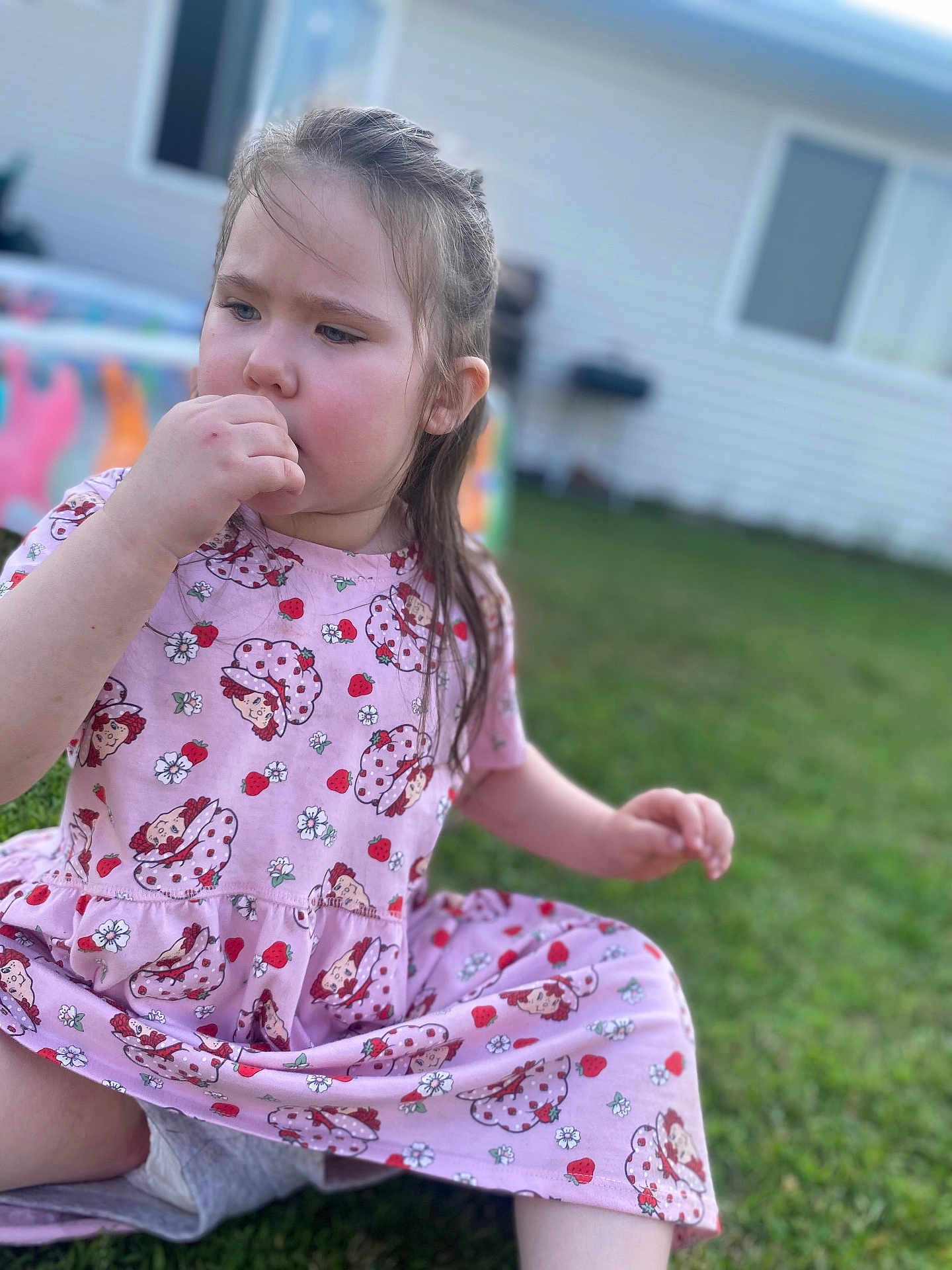 Everest Catherine Rose is registered to the contest to win money with this photo: blurred_background, cartoon_pattern, casual, child, cute, daylight, girl, grass, house, nature, outdoor, pink_dress, playtime, portrait, relaxed, sitting, strawberries, thoughtful, thumb_sucking, young