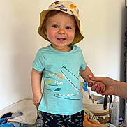 Elio a rejoint le concours — aidez-le/la à gagner de superbes lots ! toddler, child, baby, standing, bucket_hat, blue_tshirt, shorts, smiling, indoor, changing_table, hand_holding, nursery, clothing, feet, person, cute, happy, face, room, accessories