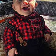 Theodore is registered to the contest to win money with this photo: baby, smiling, suspenders, shirt, red_checkered, black_pants, drool, cushion, seat, window, sunlight, indoor, happy, child, face, hand, clothing, infant, person, portrait