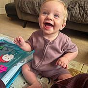 Harper is registered to the contest to win money with this photo: baby, child, smiling, happy, indoor, sitting, playmat, toy, blue_eyes, blond_hair, onesie, carpet, couch, pillow, home, person, infant, cute, cheerful, portrait