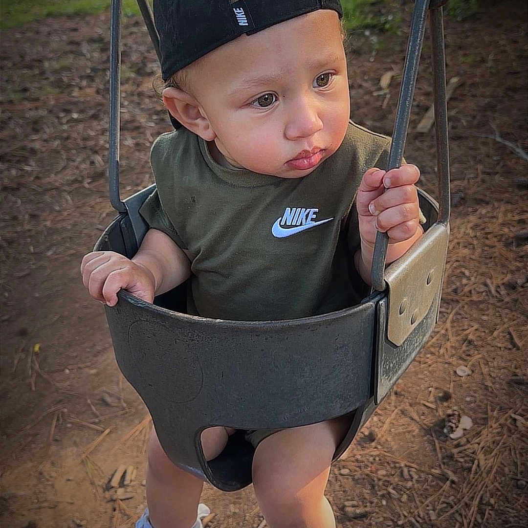 Theo is registered to the contest to win money with this photo: cap, casual_clothing, chains, child, curious, daylight, footwear, grip, nature, nike, outdoor, park, playground, seat, shirt, sneakers, swing, toddler, trees, young_child