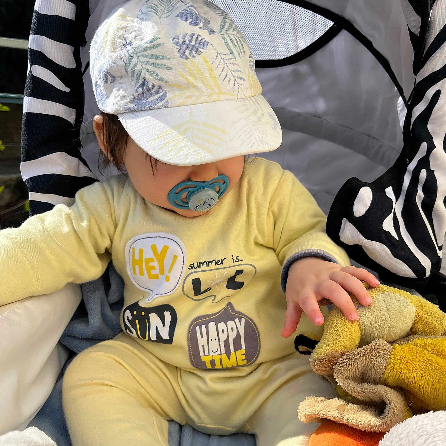 Laïon participe au concours pour gagner de l'argent avec cette photo : baby, pacifier, yellow_outfit, cap, plush_toy, stroller, zebra_pattern, child, hand, soft_toy, sunlight, comfort, cute, infant, sitting, outdoor, clothing, headwear, playful, relaxed