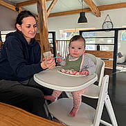 Rose a rejoint le concours — aidez-le/la à gagner de superbes lots ! baby, woman, feeding, high_chair, bib, spoon, indoor, wooden_beams, clock, window, table, floor, furniture, person, child, casual_clothing, light, home, parenting, care
